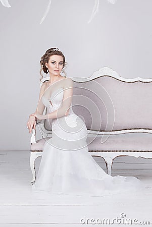 Beautiful young bride in a luxurious lace wedding dress. She sits on a white vintage sofa. Stock Photo