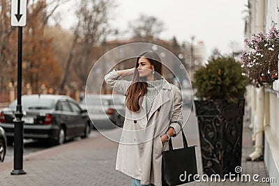 Beautiful young brown-haired woman in a sweater in a fashionable long light trench coat with a black leather bag posing on a Stock Photo