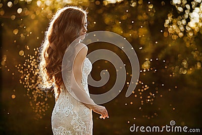 Beautiful and young brunette model girl, in white lace dress, is standing with her back at the park at the sunset Stock Photo