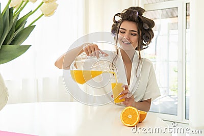 Beautiful young brunette woman in hair curlers pouring orange fresh squeezed juice from pitcher in glass sitting in Stock Photo