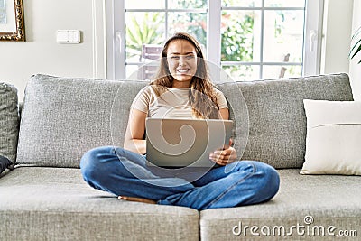 Beautiful young brunette woman sitting on the sofa using computer laptop at home with a happy and cool smile on face Stock Photo