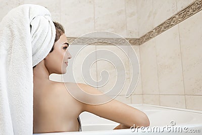 Beautiful young brunette woman wearing white towel on her head sitting in bath half a turn in bright bathroom Stock Photo