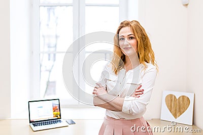 Beautiful young businesswoman standing in front of window and a desk with a laptop, crossed her arms over her chest in a white Stock Photo