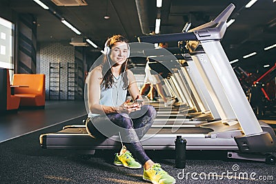 Beautiful young Caucasian girl sportswoman sitting, resting after training on treadmill against the backdrop of gym in sunny weath Stock Photo