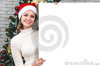 Beautiful and young caucasian woman holding and pointing to blank white board in front of big christmas tree. Concept for Stock Photo