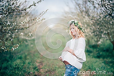 Beautiful young cheerful pregnant woman in wreath of flowers on head touching belly while walking in spring tree garden. Beauty Stock Photo