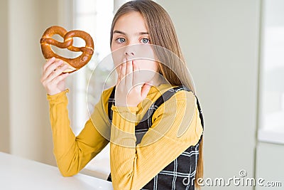 Beautiful young girl kid eating german snack salty pretzel cover mouth with hand shocked with shame for mistake, expression of Stock Photo