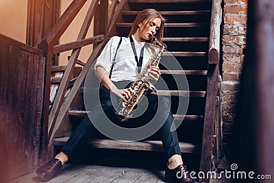 Beautiful young girl plays a saxophone sitting on steps - outdoors. Attractive woman in white shirt expression plays a saxophone Stock Photo