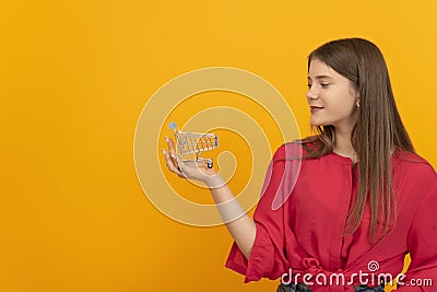 Beautiful young girl in red blouse holds grocery cart in palm of hand. Bright orange background. Buyer or customer concept. Copy Stock Photo