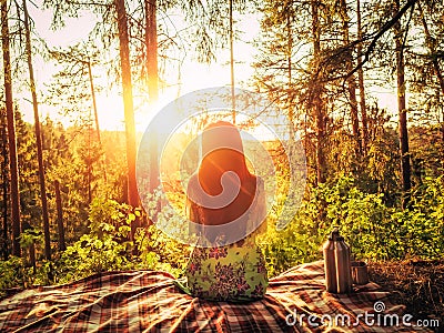 Beautiful young girl sitting on a plaid in a forest glade during sunset bright sunlight around beauty of nature. Next to Stock Photo