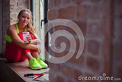 Beautiful young girl is sitting on the windowsill and drinking. Stock Photo