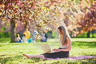 Beautiful young girl working on her laptop in park during cherry blossom season Stock Photo