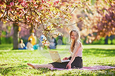Beautiful young girl working on her laptop in park during cherry blossom season Stock Photo