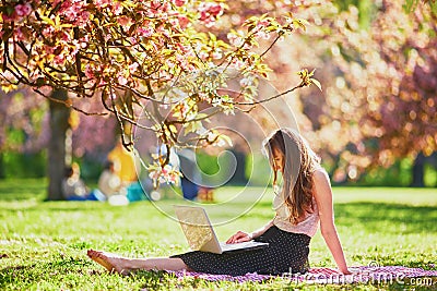 Beautiful young girl working on her laptop in park during cherry blossom season Stock Photo