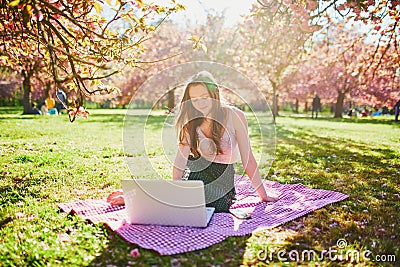 Beautiful young girl working on her laptop in park during cherry blossom season Stock Photo