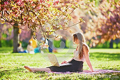 Beautiful young girl working on her laptop in park during cherry blossom season Stock Photo