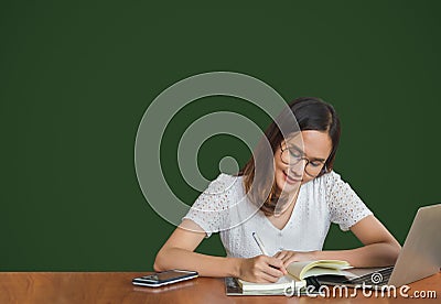 Beautiful young glasses Asian woman sitting and writing on a book with laptop at side with green background Stock Photo