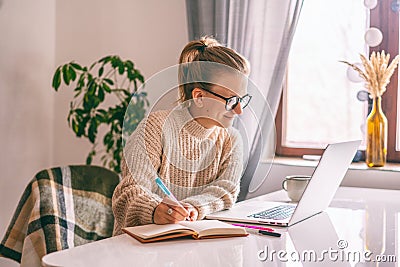 Beautiful young happy girl student with glasses and a warm siver studying at home online with a laptop writing in a notebook Stock Photo