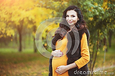 Beautiful young happy pregnant woman staying in fashion clothes in autumn park touching her belly and smiling. Stock Photo