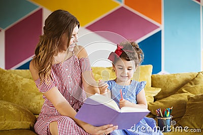 Beautiful young mother with her little cute daughter happily drawing in coloring book with felt-tip pens while spending Stock Photo