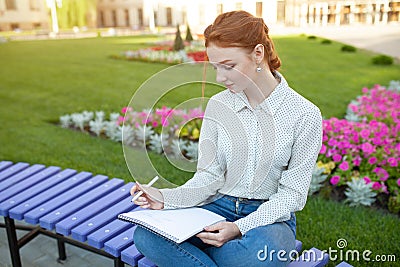 A beautiful young redhaired girl writes a romantic love letter sitting on a bench near the institute. Student love. Or homework Stock Photo