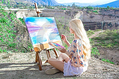 Beautiful young woman artist paints a landscape in nature. Drawing on the easel with colorful paints in the open air. Stock Photo