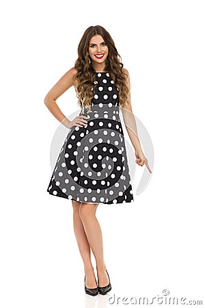 Beautiful Young Woman In Black Cocktail Dress In Polka Dots And High Heels Is Pointing Down And Smiling Stock Photo