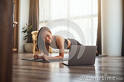 A beautiful young woman doing plank on training mat while watching online workout tutorials on laptop at home Stock Photo