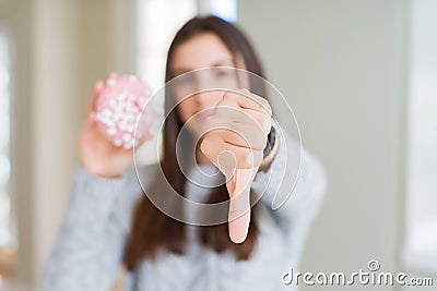 Beautiful young woman eating sugar marshmallow pink donut with angry face, negative sign showing dislike with thumbs down, Stock Photo