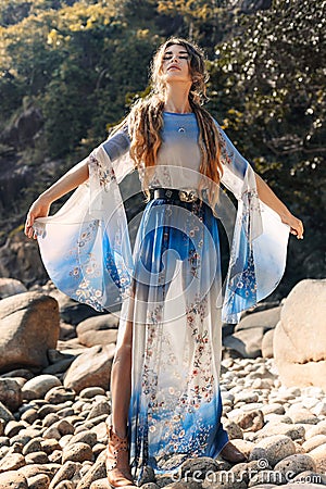 Beautiful young woman in elegant stylish dress on stone beach at sunset Stock Photo