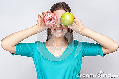 Beautiful young woman with freckles in green dress, holding before her eyes green apple and pink donut and shows her teeth. Stock Photo