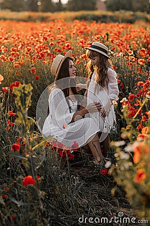 Beautiful young woman with girl in field with poppies, mother and daughter in white dresses and straw hats in evening at sunset, Stock Photo