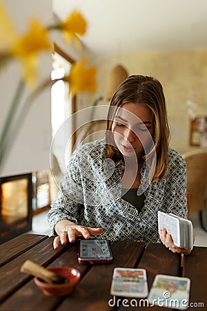 Beautiful young woman is guessing on cards with tarot, runes on wooden table and uses an online app in phone to Stock Photo