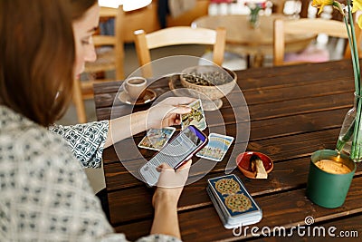 Beautiful young woman is guessing on cards with tarot, runes on wooden table and uses an online app in phone to Stock Photo