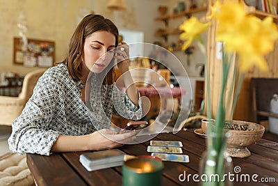 Beautiful young woman is guessing on cards with tarot, runes on wooden table and uses an online app in phone to Stock Photo