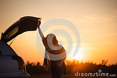 Beautiful young woman happy and dancing in a car`s trunk during a road trip in Europe in the last minutes of Golden Hour Stock Photo