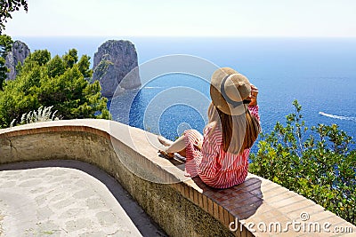 Beautiful young woman with hat sitting on wall looking at stunning panoramic view of Capri Island with Faraglioni sea stack and Stock Photo