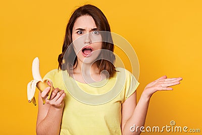 Beautiful young woman holding fresh banana, takes hands to side, has uncomprehending facial expresion, standing with open mouth Stock Photo