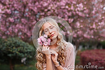 Beautiful young woman with long curly blonde hair and closed eyes holding blooming branch of sakura tree Stock Photo