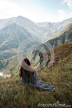 Beautiful young woman in a long dress is sitting on a cliff in the background of mountains Stock Photo