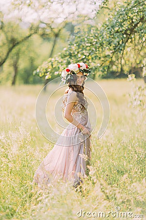 Beautiful young woman in long lilac dress with wreath on head standing back outdoors Stock Photo