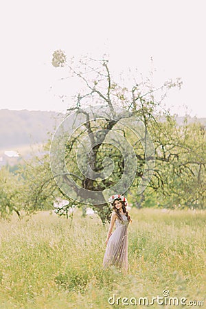 Beautiful young woman in long lilac dress with wreath on head standing near tree Stock Photo