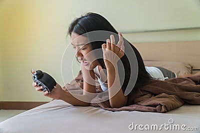 Beautiful young woman lying on a bed holding an alarm clock in her hand with exasperation Stock Photo