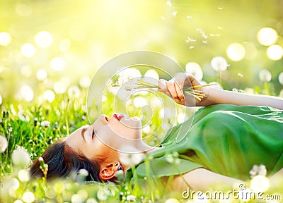 Beautiful young woman lying on the field in green grass and blowing dandelion flowers Stock Photo