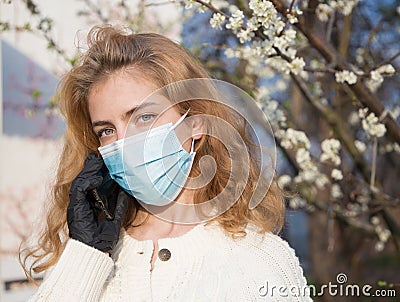 Beautiful young woman in a medical mask on the background of a blossoming spring tree speaks by phone Stock Photo