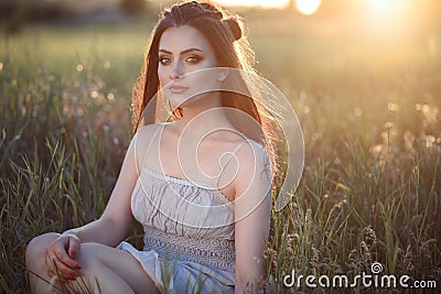 Beautiful young woman with perfect make up and long plaited hair sitting in the field at sunset Stock Photo