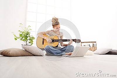 Beautiful young woman playing guitar with computer, learn to play with an online course, sitting on the floor in casual clothes Stock Photo