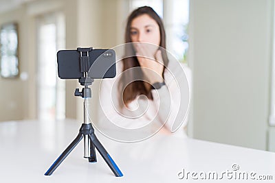 Beautiful young woman recording selfie video with smartphone webcam cover mouth with hand shocked with shame for mistake, Stock Photo