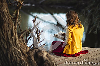 Beautiful young woman on the river banks sitting on a wooden platform with a book, teenager girl reading fiction literature on Stock Photo