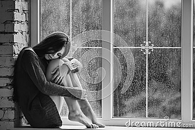 Beautiful young woman sitting alone close to window with rain drops. and sad girl. Concept of loneliness. Black Stock Photo